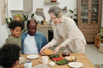Woman serving breakfast to family in cozy kitchen. Diverse family sitting around wooden table with breakfast foods, greenery in background creating warm atmosphere