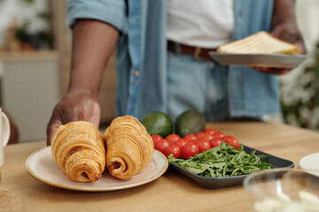 Close-up of hand serving croissants on plate with fresh avocados, cherry tomatoes, and leafy greens in background on kitchen counter