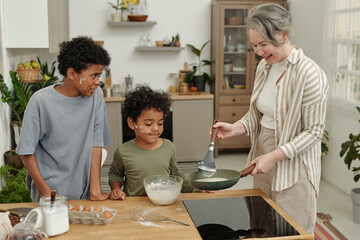 Woman and two young boys enjoying time in kitchen preparing meal together. Smiling faces and...