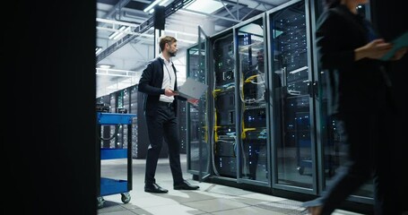 IT Technician Using Laptop Computer while a Young Female Engineer Walking Along the Server Room Corridor with a Tablet. Administrator Running Compute Diagnostics and Doing Database Maintenance Work - Powered by Adobe