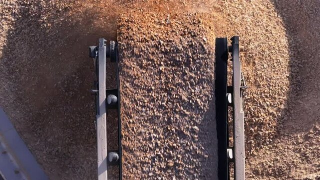 Top-down aerial view of a conveyor belt dropping crushed stone onto a large pile. Industrial machinery at a gravel pit or mining quarry for producing construction materials.
