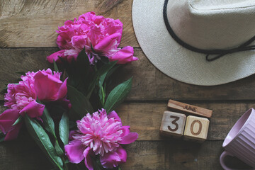 Pink peonies, straw hat and wooden calendar showing june 30th on rustic wooden table. flat lay
