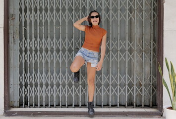 Pretty, chic Asian woman poses stylishly against a metal gate, hand on hip, in a rust crop top, distressed denim shorts, and black combat boots, looking cute with an urban edge