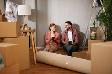 A couple sits on the floor of their new living room surrounded by unpacked boxes. They share a...