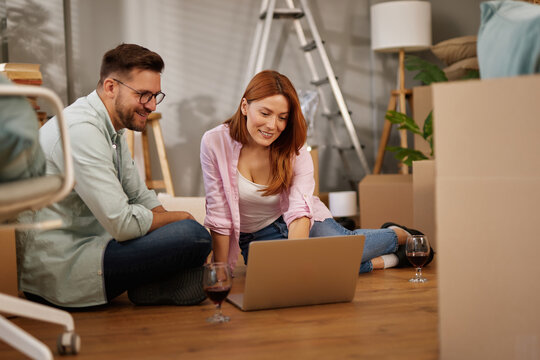 Two individuals are seated on the floor, looking at a laptop with boxes around them, enjoying each other's company while planning their new home setup.