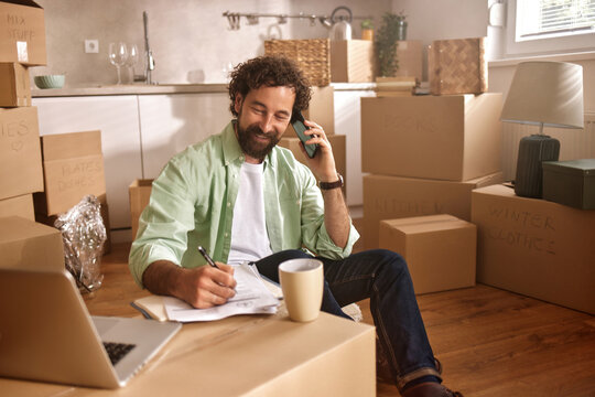A man is sitting on the floor of his new home surrounded by unpacked boxes. He is talking on the phone while taking notes, showcasing his moving preparations. - Powered by Adobe