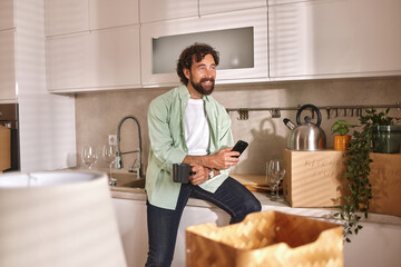 A man smiles while holding a cup of coffee and phone in a kitchen filled with moving boxes. He is...