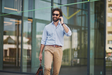A man in business attire walks with a briefcase while engaged in a phone conversation outside a...