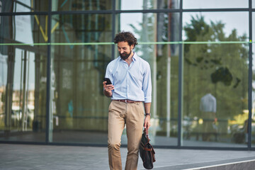 A business professional dressed in a shirt and tan trousers checks his smartphone while walking...