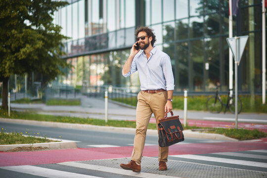 A business professional strides across a pedestrian crosswalk, engaged in a phone call. The modern urban environment showcases sleek buildings and green landscaping.