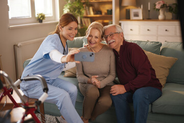 A young female home health aide in scrubs sits with a smiling elderly couple on their sofa and...