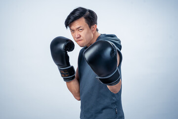 Asian man in sleeveless hoodie wearing boxing gloves strikes forward fighting pose with intense...