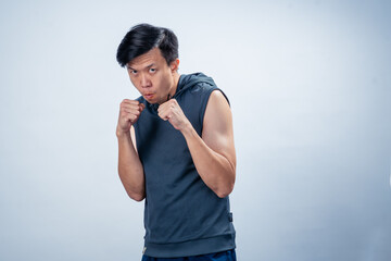 Asian man wearing sleeveless hoodie strikes boxing stance with clenched fists and focused face,...