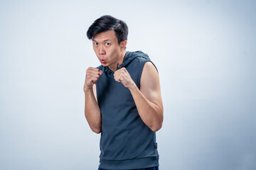 Asian man wearing sleeveless hoodie strikes boxing stance with clenched fists and focused face,...