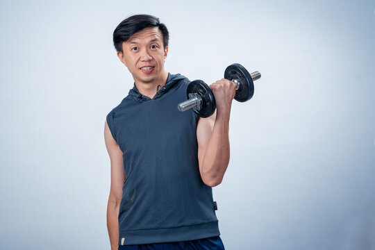 Asian man in sleeveless hoodie lifting a dumbbell with one arm and smiling, posing confidently in studio with plain light background, showing strength, motivation, and fitness commitment
