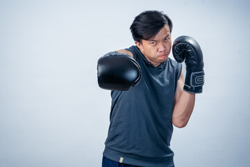 Asian man in sleeveless hoodie wearing boxing gloves throws forward punch with intense facial...