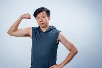 Asian man in sleeveless hoodie flexes arm and makes straight face while posing confidently in front...