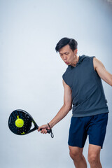 Asian man in sleeveless hoodie and shorts holding padel racket prepares for swing with focused face...