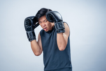 Asian man in sleeveless hoodie wearing boxing gloves strikes forward fighting pose with intense expression, ready to attack in studio with plain light background, showing strength and focus