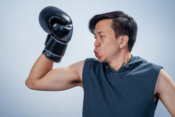 Asian man in sleeveless hoodie wearing black boxing glove raises fist and smiles widely, posing...