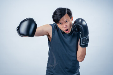 Asian man in sleeveless hoodie wearing boxing gloves throws forward punch with intense facial...