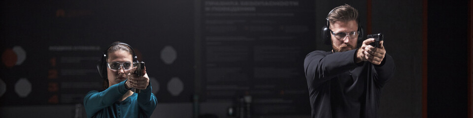Header of young Caucasian woman and man practicing handgun shooting at indoor shooting club, both...
