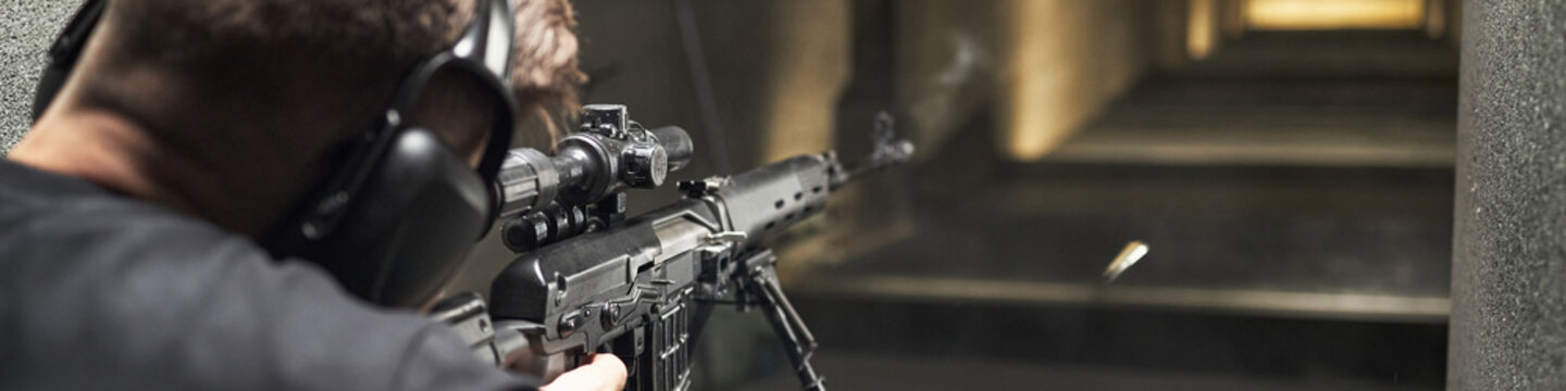 Header of man aiming rifle with scope at indoor range, wearing protective earmuffs, bullet captured mid-flight, participating in shooting club activity