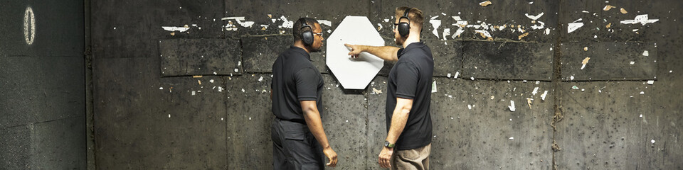 Caucasian shooting coach instructing and Black middle aged man standing at indoor range, wearing...