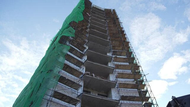 Incomplete office tower wears ragged tarps flapping noisily in strong wind. Workers in hard hats move cautiously across metal frameworks