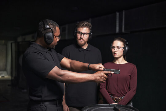 African American man instructing Caucasian young adult man and woman at indoor shooting club, all wearing protective earmuffs and safety glasses, focusing on handgun handling