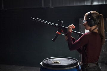 Fototapeta premium Young woman wearing protective goggles and earmuffs loading semi automatic rifle at indoor shooting club range, standing beside blue barrel with ammunition visible on top