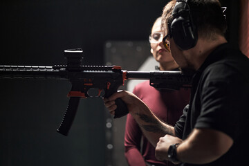 Man wearing protective earmuffs instructing Caucasian young woman on holding rifle at shooting club indoor range, both focusing on firearm handling