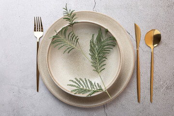 Top view of beige ceramic plates with fern leaf and golden cutlery on a textured cracked stone...