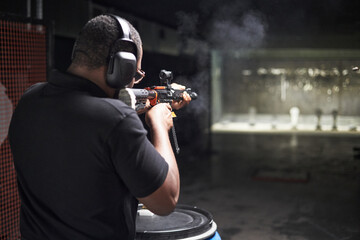 Black man wearing protective earmuffs aiming and firing rifle at shooting range targets during practice session, standing with focused posture and holding firearm with both hands