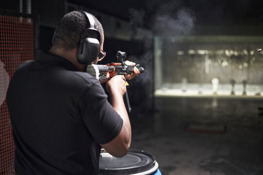 Black man wearing protective earmuffs aiming and firing rifle at shooting range targets during practice session, standing with focused posture and holding firearm with both hands