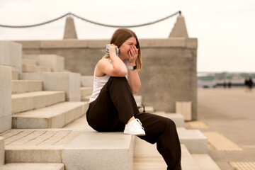 Young woman sitting on stone steps, laughing while talking on phone outdoors. Natural feel with...