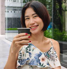 Beautiful Asian woman in a stylish floral dress holds a paper cup of coffee, embracing an eco-conscious urban lifestyle