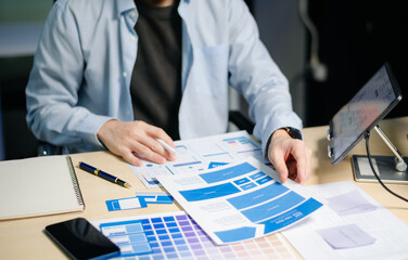 Asian designer’s hands reviewing UX/UI mockups and color palettes at modern desk. App design,...