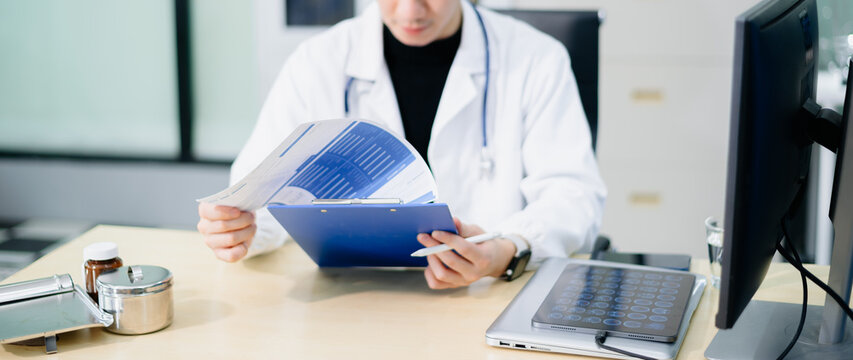 Asian doctor in white coat reviewing medical report on clipboard at office desk with stethoscope and tablet. - Powered by Adobe