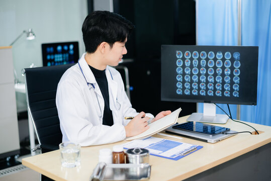 Asian doctor studying brain MRI scans on monitor and writing notes in modern clinic. Concept of radiology, neurology