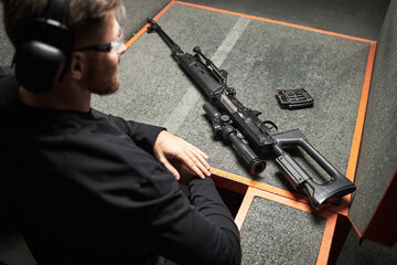 Caucasian young man wearing protective earmuffs sitting at shooting range table preparing to use...