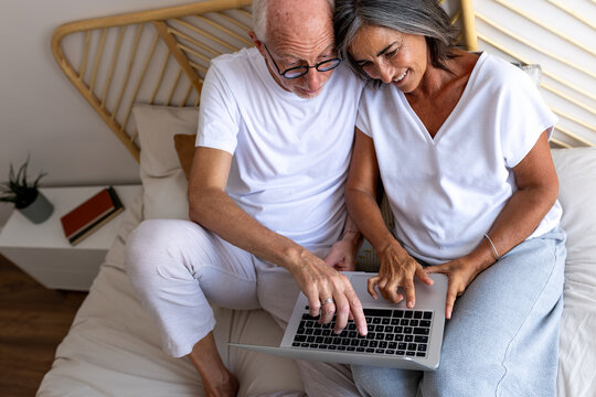 High angle view of mature caucasian couple using laptop together relaxing sitting on bed. Man pointing finger to screen - Powered by Adobe