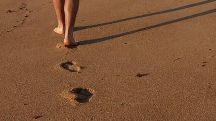 Follow shot of a Barefoot male walking to the camera leisurely along golden sandy shoreline, leaving distinctive footprints behind while reflecting peaceful seaside moments during summer vacation - Powered by Adobe