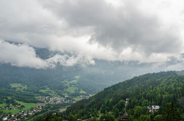 Mountain peaks covered in snow and clouds with green forest trees in foreground, dramatic sky