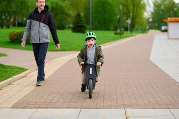 child riding scooter in park while dad walks nearby. enjoyable father-son outdoor activity on bright day. emphasizes family bonding, youth adventure, active lifestyle, springtime bliss