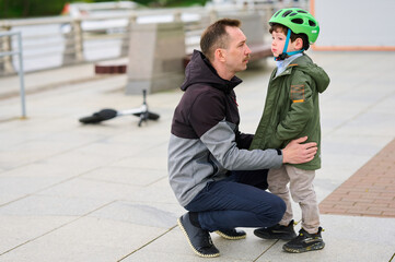 father comforts son wearing green helmet after scooter fall on paved city walkway. emotional support, parenting, and outdoor safety emphasized in family-oriented urban scene