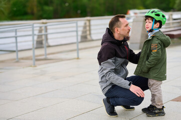 father kneeling with son wearing helmet on sidewalk by railing outdoors. parent-child relationship...