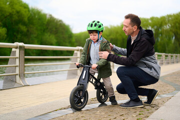 dad teaching son to ride balance bike on park pathway, green helmet for safety, supportive bonding...