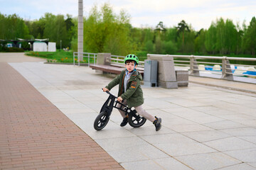 young boy riding balance bike on urban walkway during spring. wearing green helmet and jacket, enjoying outdoor adventure and learning new skills. greenery and benches in background