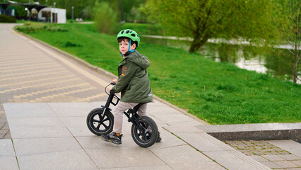 child on balance bike wearing helmet looks over shoulder in park. young rider explores outdoors,...